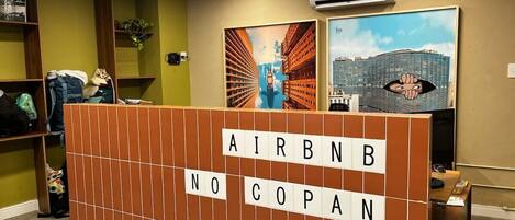 Un espace d’accueil avec une cloison en bois sur laquelle est inscrit « AIRBNB NO COPAN », et deux œuvres d’art encadrées accrochées au mur.