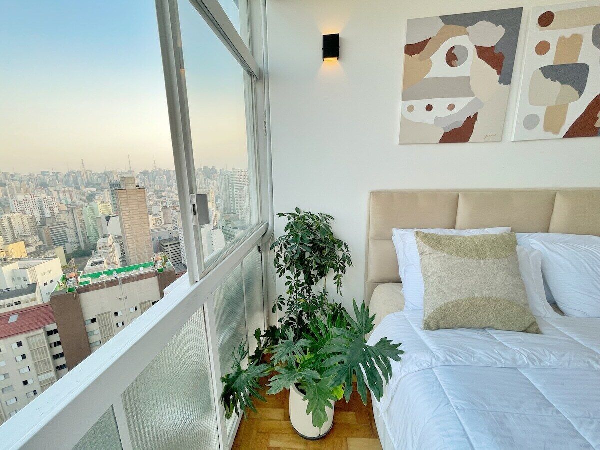 A bedroom with a city view, a bed with white bedding, a potted plant, and abstract wall art.
