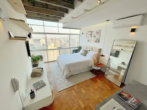A modern bedroom with a large bed, a desk, a mirror, and a city view. - 2707B (São Paulo)