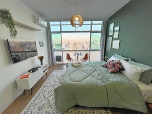 A modern bedroom with a large bed, a TV mounted on the wall, a small table and chairs by the window, and a view of the cityscape. - 2709B (São Paulo)