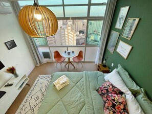 Ein Hotelzimmer mit großem Fenster, einem Bett mit grüner Bettdecke, einem kleinen Tisch mit Stühlen und Bildern an den Wänden.