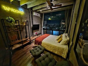 Una habitación con una cama, un estante con objetos, un letrero de neón y vista a la ciudad de noche.