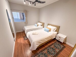 Ein Schlafzimmer mit zwei Betten, einem fensterbelaubten Fenster, einem Deckenventilator und einem Nachttisch mit Schublade.