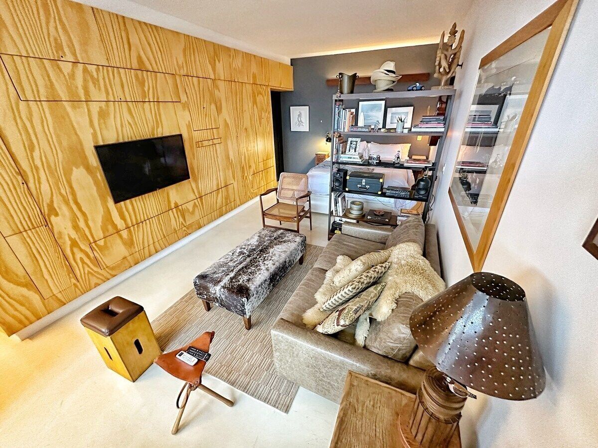 A modern living room with a wooden wall panel, a flat-screen TV, a sofa with patterned cushions, a small wooden stool, and a floor lamp.