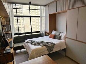 Ein modernes Schlafzimmer mit Bett, Nachttisch, Lampe und Blick auf Gebäude durch große Fenster.