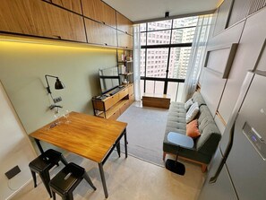 A compact living space with a kitchenette, a dining area, a sofa, and a large window providing a view of the cityscape. - 1305B (São Paulo)