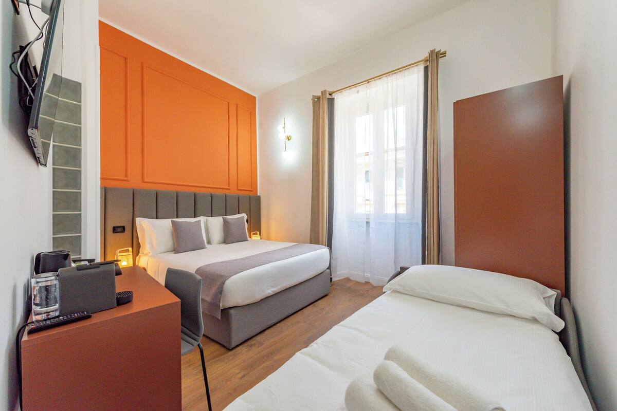 Une chambre d’hôtel avec un lit, un bureau avec un ordinateur portable, une chaise, un téléviseur fixé au mur et une fenêtre avec des rideaux.
