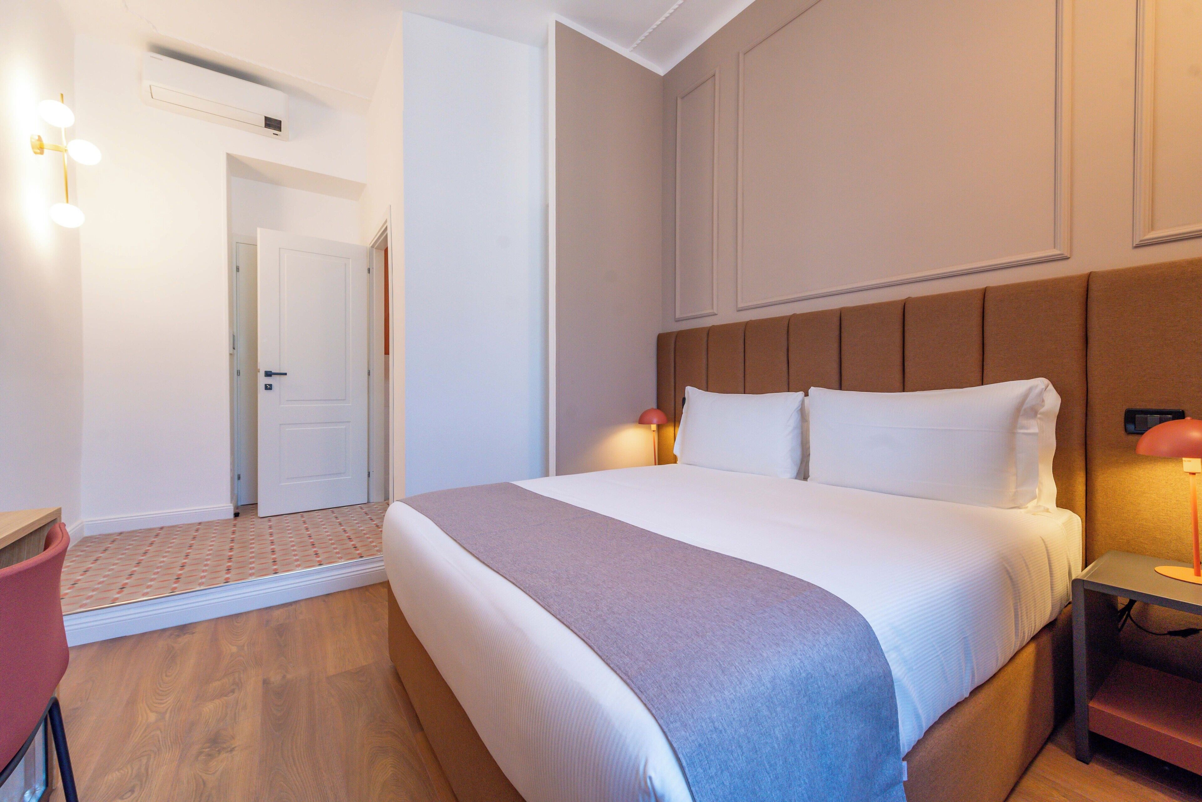 A hotel room with a bed, bedside table, lamp, and a door leading to another room.
