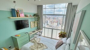 Een moderne slaapkamer met een bed, een bureau, een televisie en een groot raam met uitzicht op de stad.