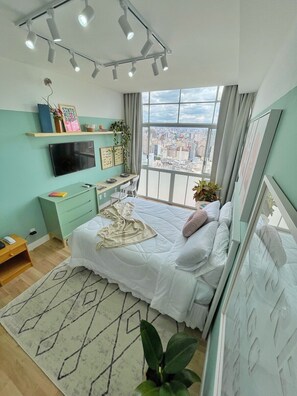 A modern bedroom with a bed, a desk, a TV, and a large window offering a city view. - 3003B (São Paulo)