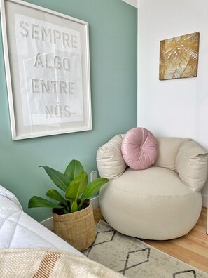 Ein Zimmer mit grüner Wand, einem gerahmten Motivationsspruch, einem runden beigefarbenen Sofa mit rossem Kissen, einer Topfpflanze und einem Teppich mit geometrischem Muster.