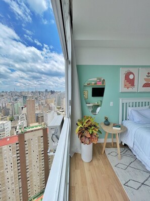 A bedroom with a city view, a round mirror, a small table with a plant, and a bed with white bedding. - 3003B (São Paulo)