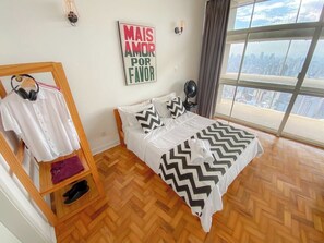 A bedroom with a bed, a wooden shoe rack, a wall-mounted light, a city view, and a motivational poster. - 2919B (São Paulo)