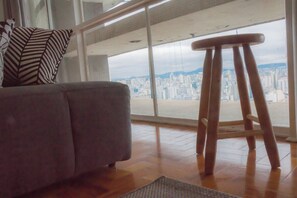 A living room with a grey sofa, a wooden stool, and a large window offering a city view. - 2919B (São Paulo)
