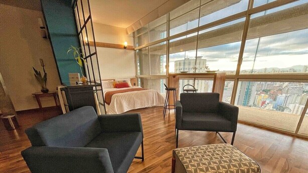 Een moderne hotelkamer met een groot raam, een bed, twee fauteuils, een bijzettafeltje en een plant in pot.