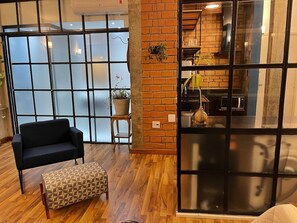 A room with a black sofa, a patterned ottoman, a wooden floor, a brick wall, and a sliding glass door leading to a kitchen area. - 2716B (São Paulo)