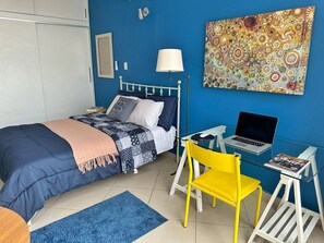 Ein Schlafzimmer mit einem Bett, einem Schreibtisch mit Laptop, einem gelben Sessel und einem blauen Teppich.