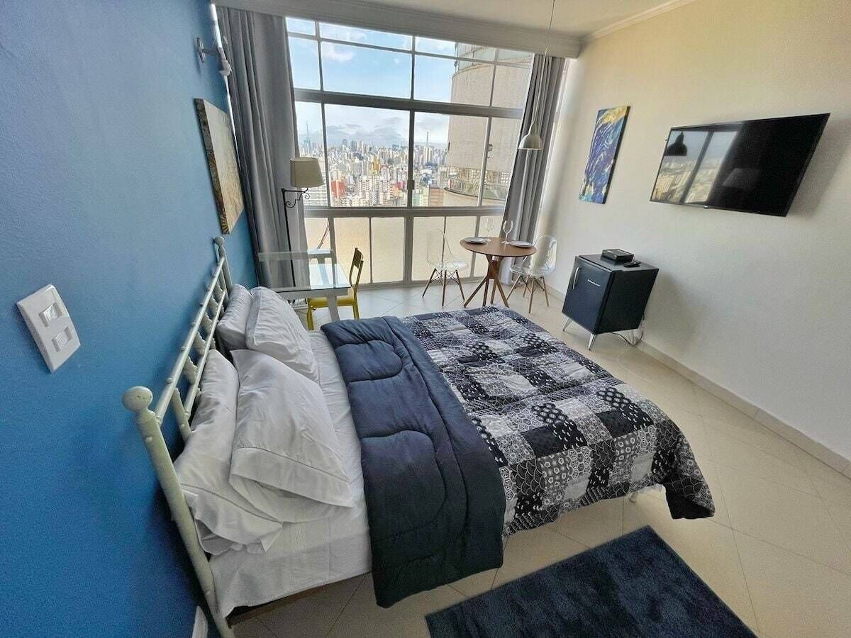 Une chambre à coucher comprenant un lit, un bureau, une chaise, un téléviseur et une vue sur la ville grâce à une grande fenêtre.