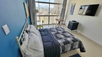 Une chambre à coucher comprenant un lit, un bureau, une chaise, un téléviseur et une vue sur la ville grâce à une grande fenêtre.