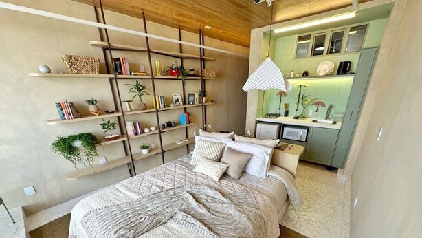 A bedroom with a bed, bedside tables, a bookshelf, a hanging light, and a kitchenette in the background. - 3206B (São Paulo)