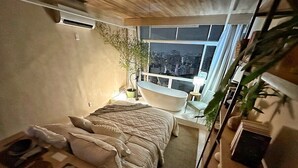 A compact bedroom with a bed, a small table, a window with a city view, and a bathtub. - 3206B (São Paulo)