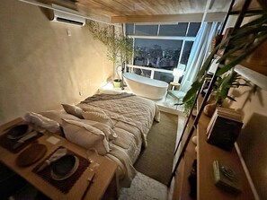 Ein gemütliches Schlafzimmer mit Bett, kleinem Tisch, Fenster mit Blick auf die Stadt und einer Badewanne.