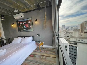 A hotel room with a bed, a desk, a chair, a window with a city view, and a hanging light fixture. - 2006B (São Paulo)