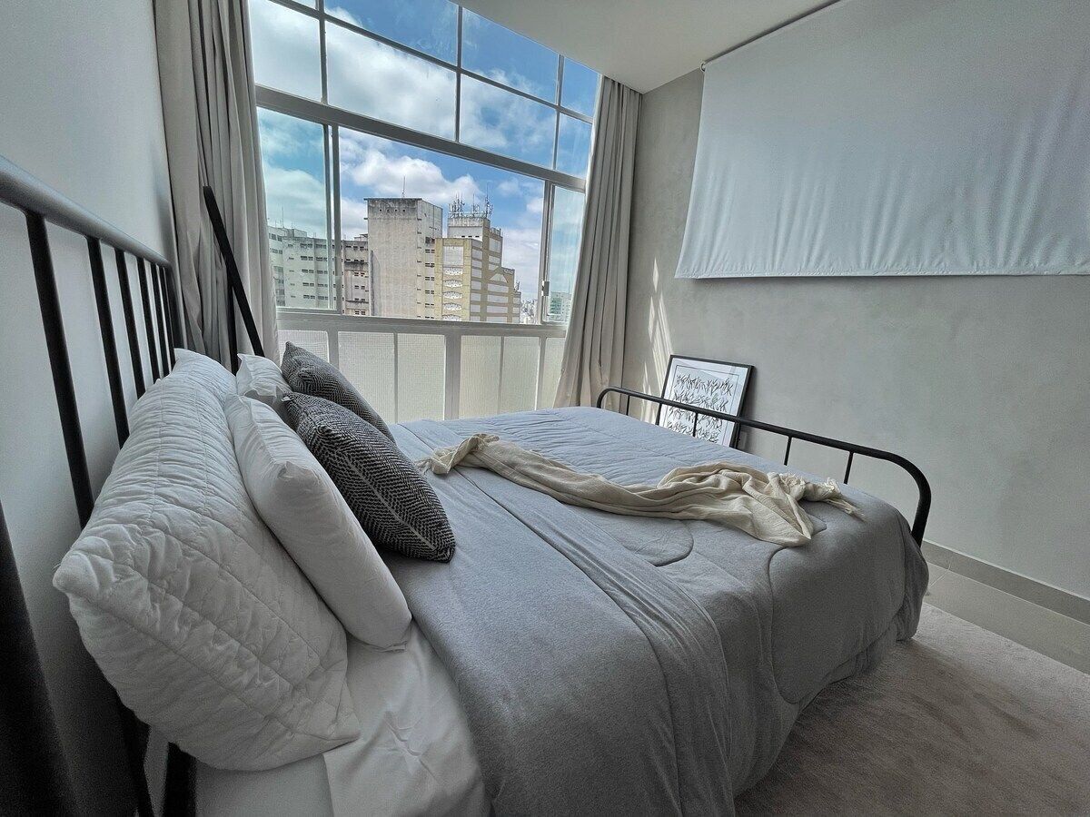 A bedroom with a bed, a window with curtains, and a view of buildings.