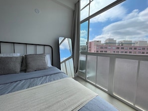 A bedroom with a large window, a bed with grey bedding, and a view of buildings outside. - 1902B (São Paulo)