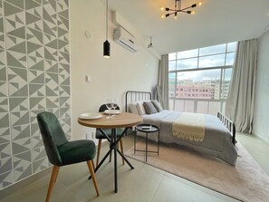 A modern bedroom with a bed, a round dining table, and a chair. There is a window with a view of buildings outside. - 1902B (São Paulo)