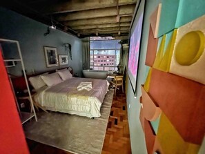 A room with a bed, a small table, a chair, and a window with a view of buildings. - 2002B (São Paulo)