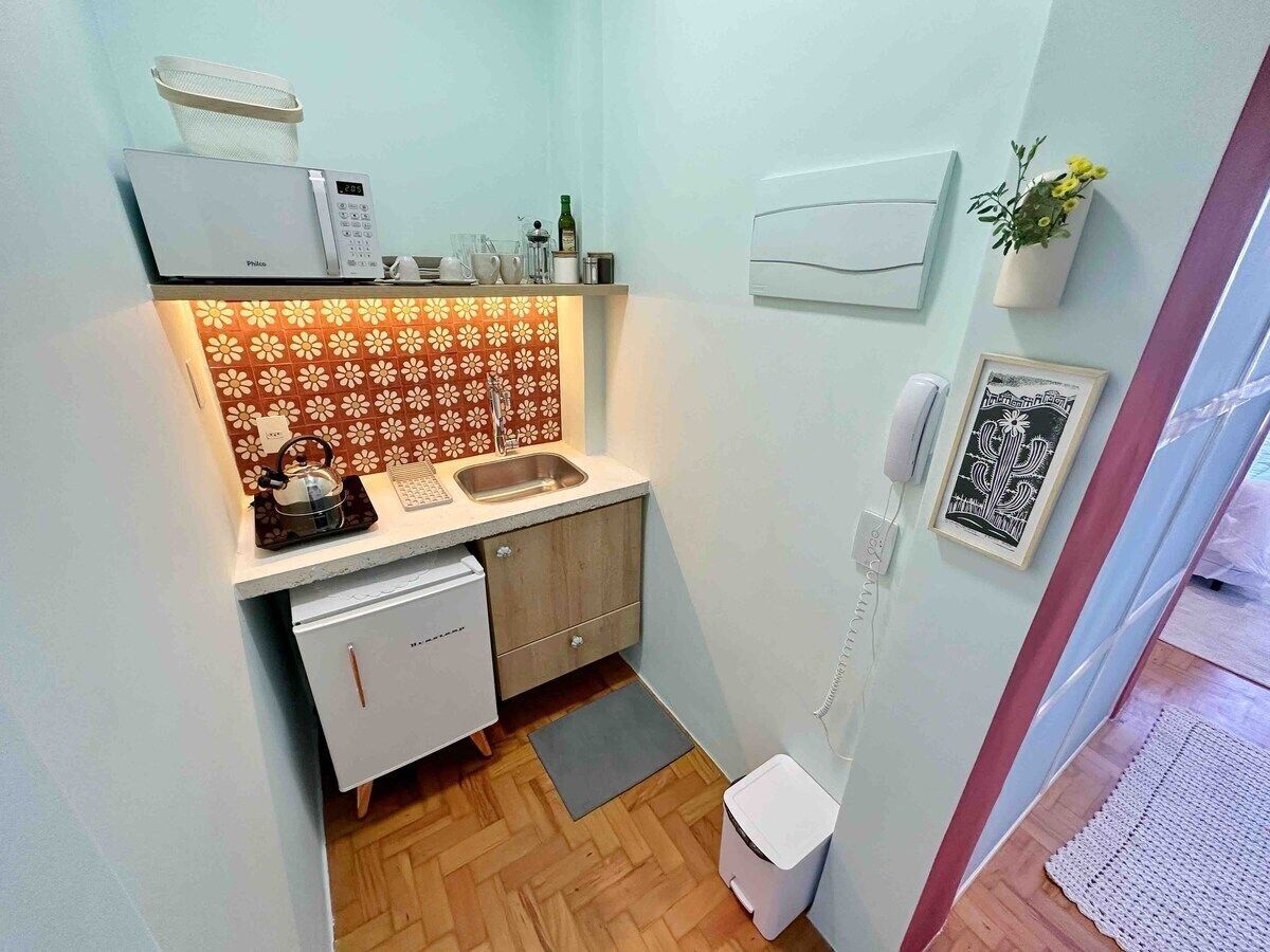 A compact kitchen with a dishwasher, a coffee machine, and a sink. There is a small refrigerator, a potted plant, and a framed picture on the wall.