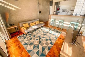 Ein kompakter Wohnbereich mit einer Couch, einer Essecke mit Hockern, einer Küchenzeile und einem gemusterten Teppich.