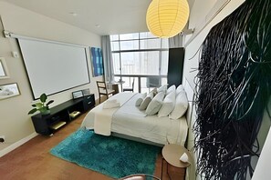 Ein modernes Schlafzimmer mit Bett, Schreibtisch, Stuhl, großem Fenster und Projektionsfläche.