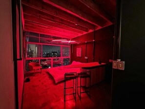 Une chambre avec un lit, une fenêtre donnant sur la ville et un plafond éclairé de lumière rouge.