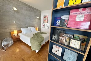 A hotel room with a bed, bedside lamps, a fan, and a bookshelf with various items. - 0818B (São Paulo)