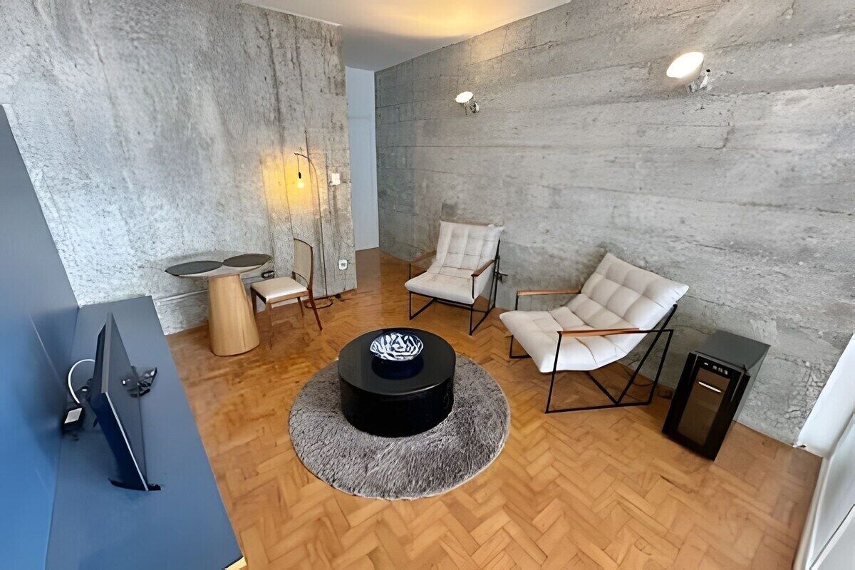 Ein modernes Wohnzimmer mit Fischgrätparkett, einem runden schwarzen Couchtisch, zwei weißen Ledersesseln, einem kleinen Holznachttisch und einer schwarzen TV-Konsole.