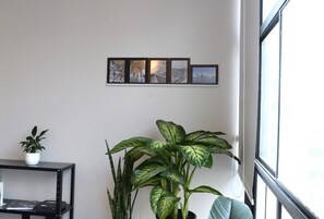 A room with a large window, a black metal shelf with a potted plant, and a wall-mounted art piece. - 1207B (São Paulo)