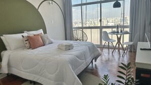 A bedroom with a large bed, a dining table with chairs, a view of the city through large windows, and a potted plant. - 2606B (São Paulo)