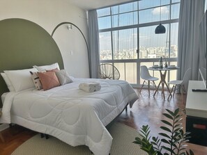 A bedroom with a large bed, a dining table with chairs, a view of the city through large windows, and a potted plant. - 2606B (São Paulo)