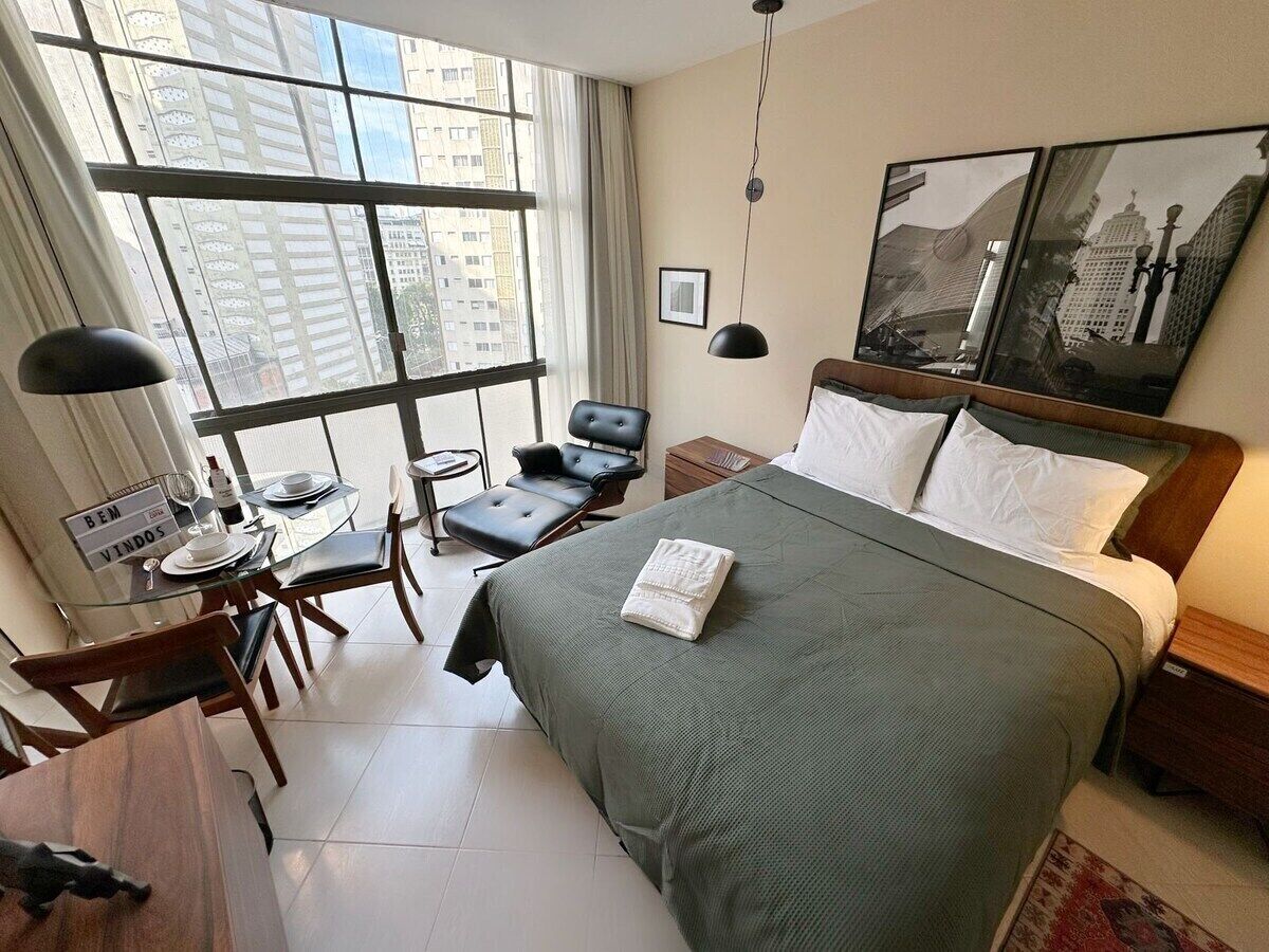 Ein Hotelzimmer mit einem großen Bett, einem Schreibtisch mit Stuhl, einem kleinen Tisch und einem Fenster mit Blick auf Gebäude.