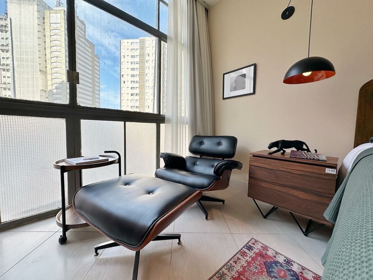 A modern bedroom with a large window, a black leather chair, a wooden dresser, and a small table.