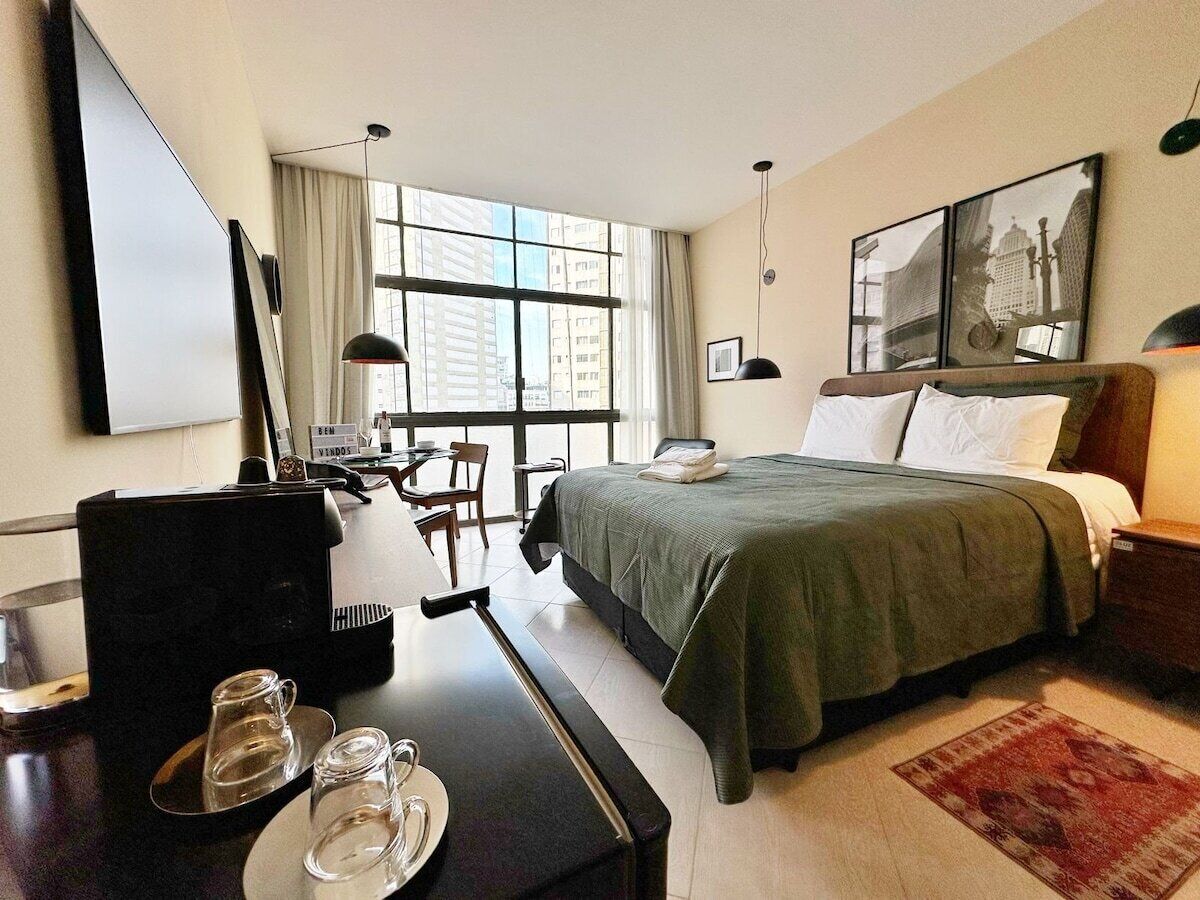 A hotel room with a bed, a desk with a computer, a coffee machine, and a window with a city view.
