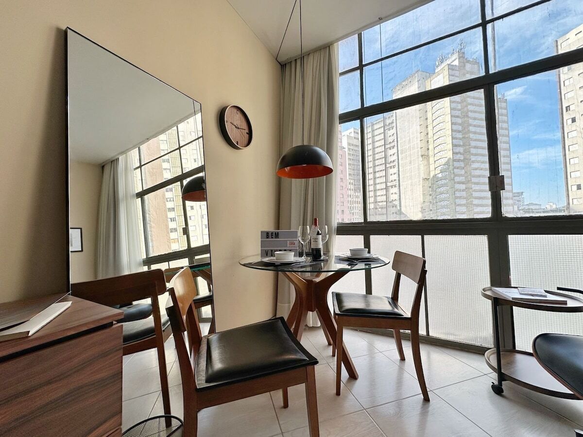 Quarto de hotel com uma janela grande, uma mesa redonda, duas cadeiras, uma escrivaninha, um espelho, um relógio e uma placa com a palavra 'BEM'.