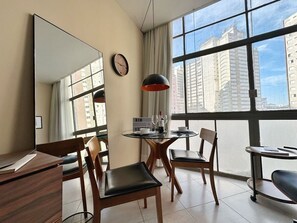 Ein Hotelzimmer mit großem Fenster, einem runden Tisch, zwei Stühlen, einem Schreibtisch, einem Spiegel, einer Uhr und einem Schild mit der Aufschrift 'BEM'.