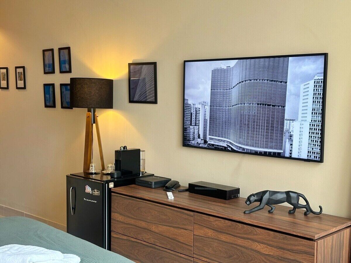 Quarto de hotel com cama, escrivaninha com luminária, geladeira, TV de tela plana e uma imagem da paisagem urbana na parede.