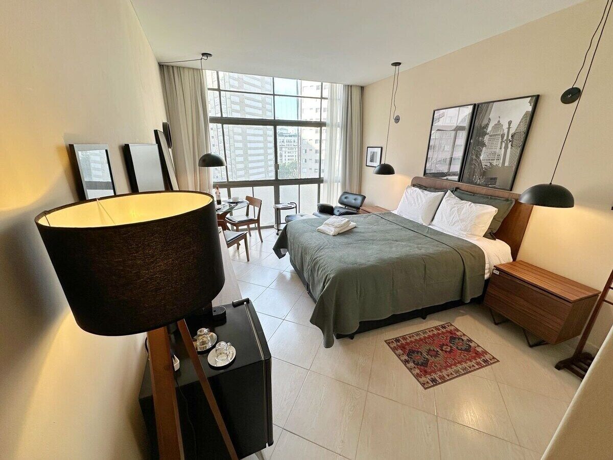 A hotel room with a bed, bedside table, lamp, and a view of buildings through the window.