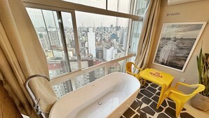 Une salle de bain avec une baignoire, une fenêtre donnant sur la ville, une table jaune et une plante en pot.
