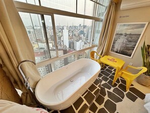 A bathroom with a bathtub, a window with a city view, a yellow table, and a potted plant. - 2610B (São Paulo)