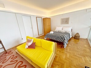 Ein Zimmer mit gelbem Sofa, einem Bett mit geometrischem Bettbezug und einem hölzernen Kleiderschrank.